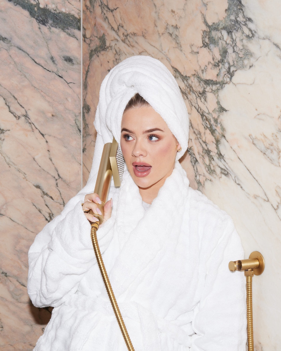 A woman in a white bathrobe and head towel holding a gold handheld shower filter